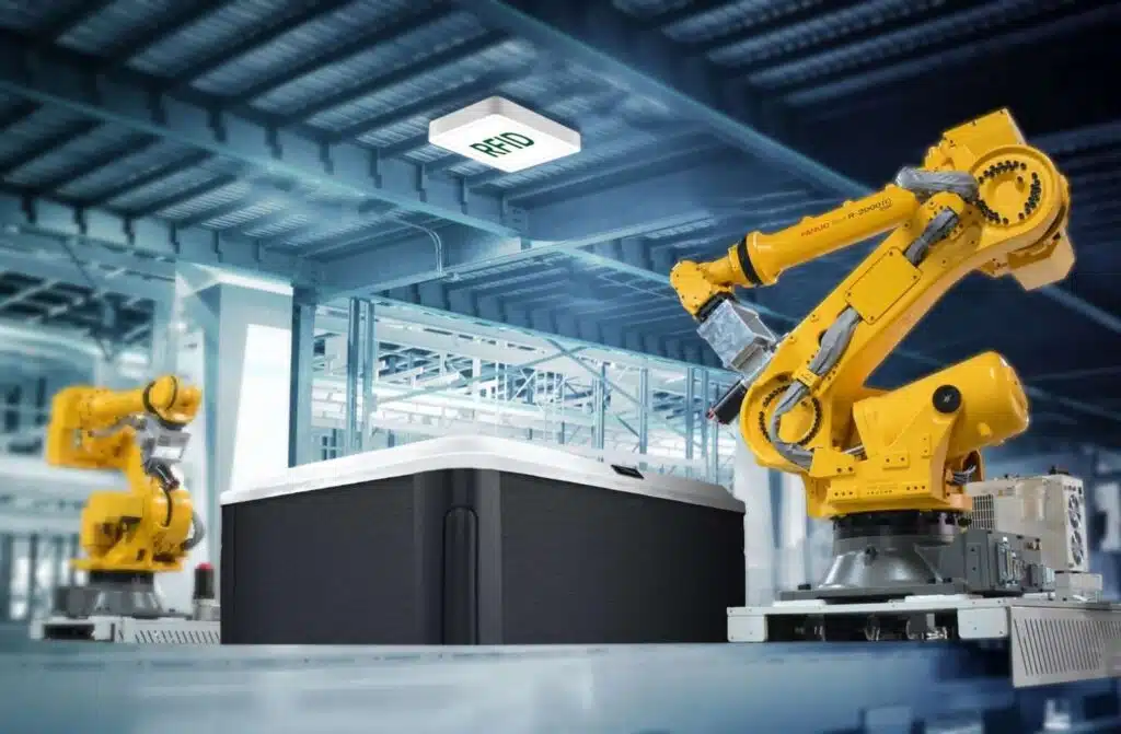 Deux bras robotisés industriels jaunes travaillent avec un grand conteneur dans une usine moderne, avec un capteur RFID monté au plafond au-dessus d'eux - une technologie souvent utilisée dans l'automatisation de la fabrication de spas et de jacuzzis.