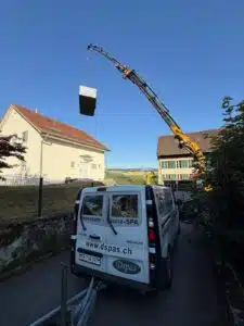 Une grue soulève un grand objet rectangulaire au-dessus d'une maison. Une camionnette blanche portant les inscriptions "www.dspas.ch" et "Hammam - Sauna - SPA" est garée dans une rue étroite à côté de la grue. Le ciel est clair et bleu.