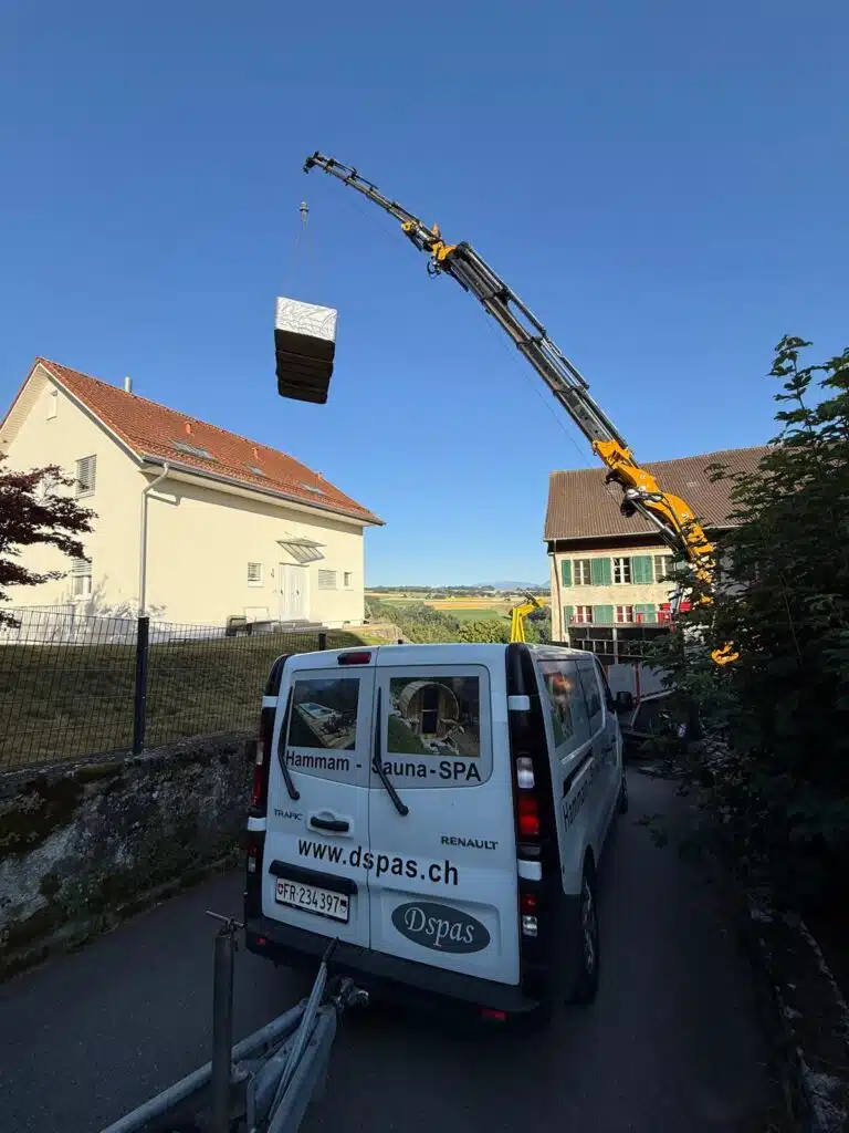 Une grue soulève un grand objet rectangulaire au-dessus d'une maison. Une camionnette blanche portant les inscriptions "www.dspas.ch" et "Hammam - Sauna - SPA" est garée dans une rue étroite à côté de la grue. Le ciel est clair et bleu.