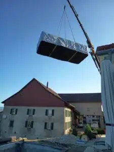 Une grue soulève un matelas enveloppé dans du plastique au-dessus d'une maison dans un ciel bleu clair, avec d'autres bâtiments et un jardin visibles en contrebas.
