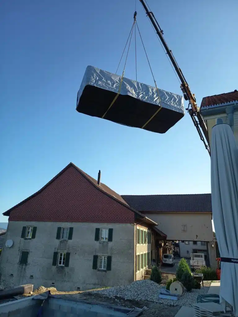 Une grue soulève un matelas enveloppé dans du plastique au-dessus d'une maison dans un ciel bleu clair, avec d'autres bâtiments et un jardin visibles en contrebas.