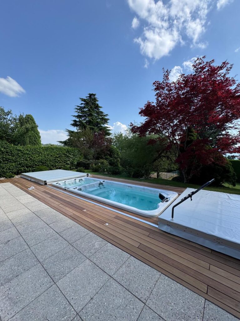 Une petite piscine rectangulaire à l'eau claire se trouve sur une terrasse entourée de verdure, d'arbres et d'un ciel bleu nuageux. L'endroit est paisible et bien entretenu, les couvertures de piscine ayant été partiellement enlevées.