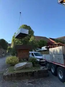 Un grand objet enveloppé d'un isolant argenté est soulevé par une grue au-dessus d'une rue de village, près d'un panneau en bois et d'un puits. Un camion et une camionnette sont garés à proximité, sous un ciel dégagé.