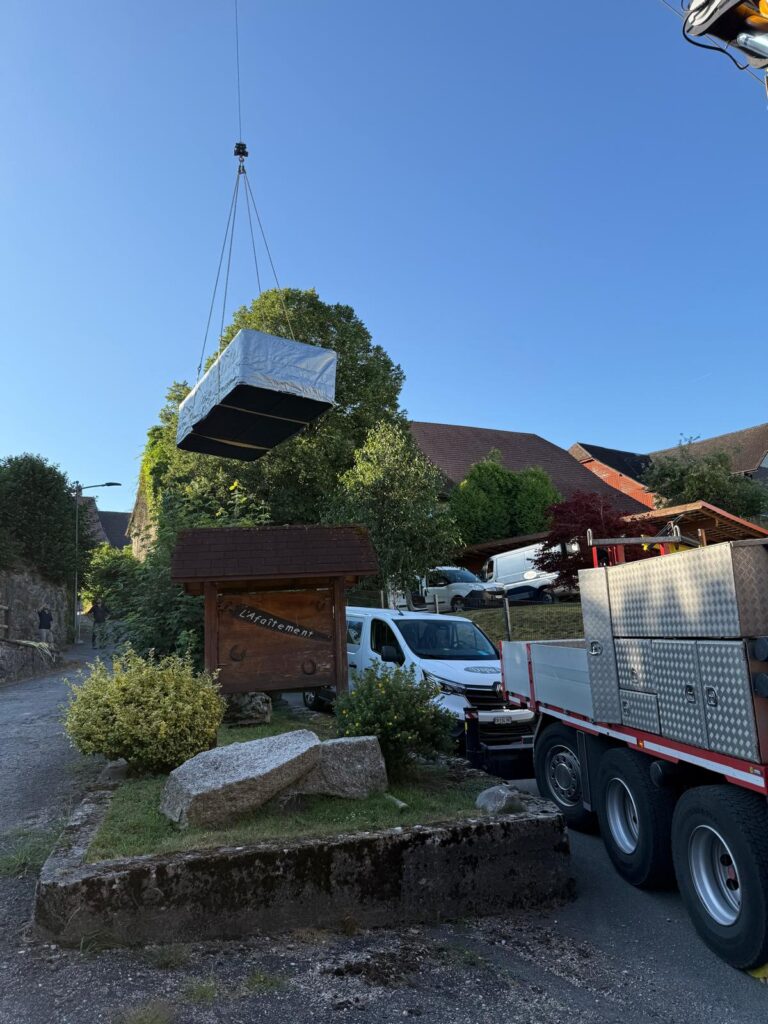 Un grand objet enveloppé d'un isolant argenté est soulevé par une grue au-dessus d'une rue de village, près d'un panneau en bois et d'un puits. Un camion et une camionnette sont garés à proximité, sous un ciel dégagé.