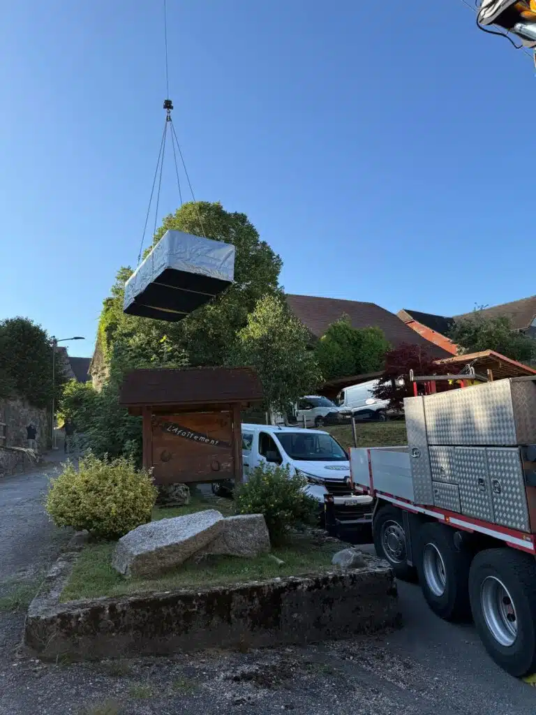 Un grand objet enveloppé d'un isolant argenté est soulevé par une grue au-dessus d'une rue de village, près d'un panneau en bois et d'un puits. Un camion et une camionnette sont garés à proximité, sous un ciel dégagé.