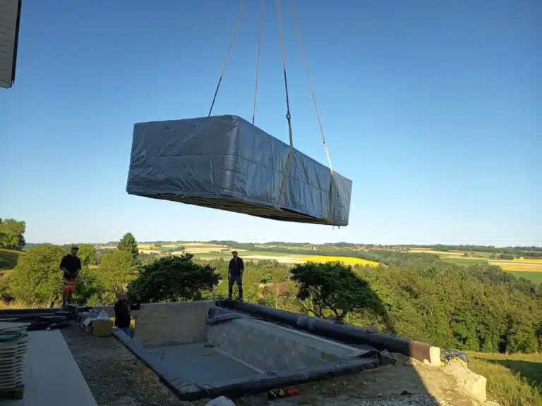 Une grande piscine rectangulaire, enveloppée d'un matériau de protection, est soulevée par une grue au-dessus d'un chantier de construction, des ouvriers se tenant à proximité. Le paysage à l'arrière-plan est composé de champs et d'arbres sous un ciel bleu clair.