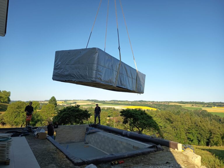 Une grande piscine rectangulaire enveloppée d'un matériau de protection est soulevée par une grue au-dessus d'une fosse vide. Trois personnes se tiennent à proximité et un paysage pittoresque se dessine à l'arrière-plan.