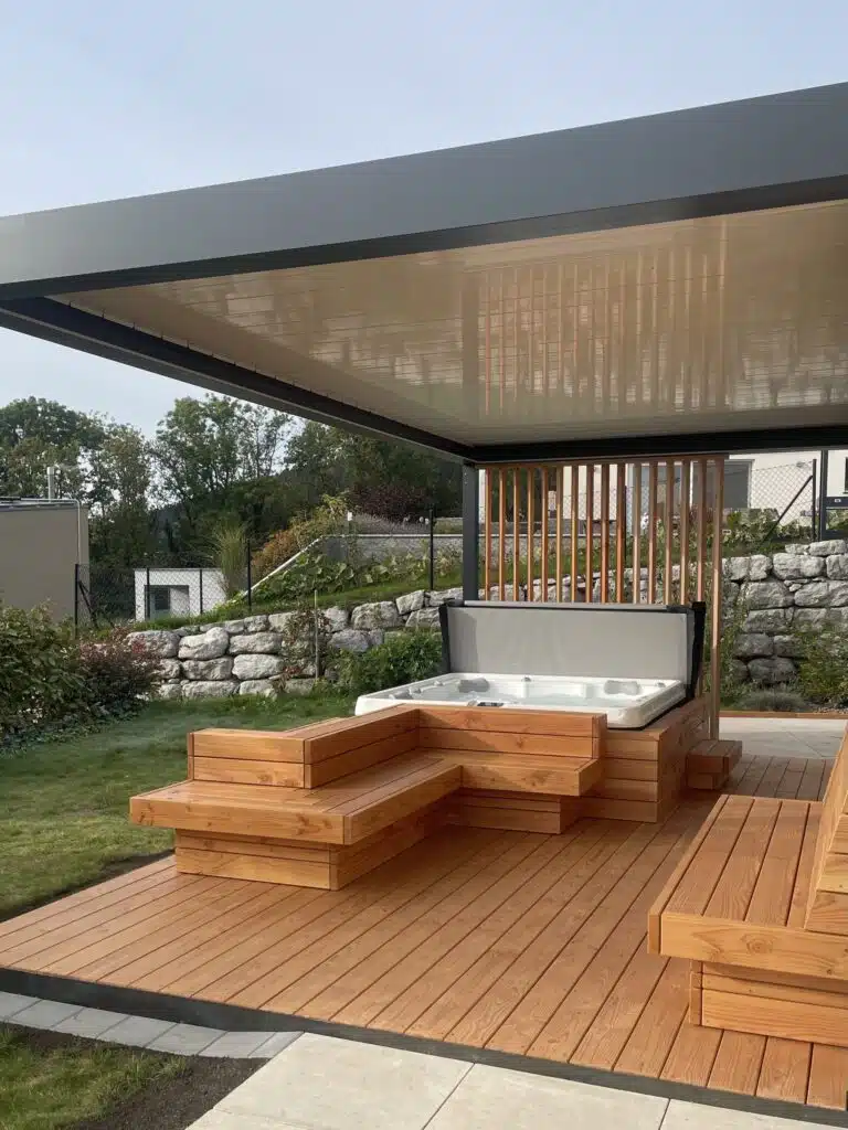 Un patio couvert moderne avec une terrasse en bois comprend un jacuzzi intégré et des bancs. La zone est entourée de verdure et d'un aménagement paysager en pierre, créant ainsi un espace extérieur relaxant.
