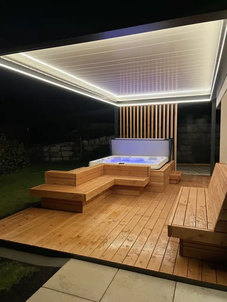 Un spa extérieur moderne avec une terrasse en bois, des sièges intégrés et un éclairage LED en hauteur, abrité par un élégant toit blanc la nuit. L'endroit est élégant, propre et accueillant.