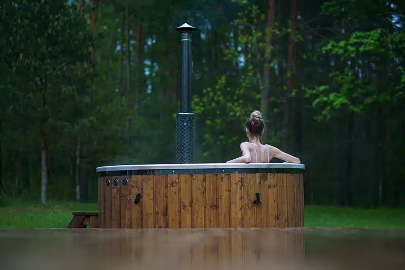 Une personne aux cheveux blonds coiffés en chignon se détend seule dans un jacuzzi rond en bois en plein air, profitant de l'atmosphère calme et paisible d'un bain nordique suisse au milieu d'arbres verts et d'une forêt luxuriante.