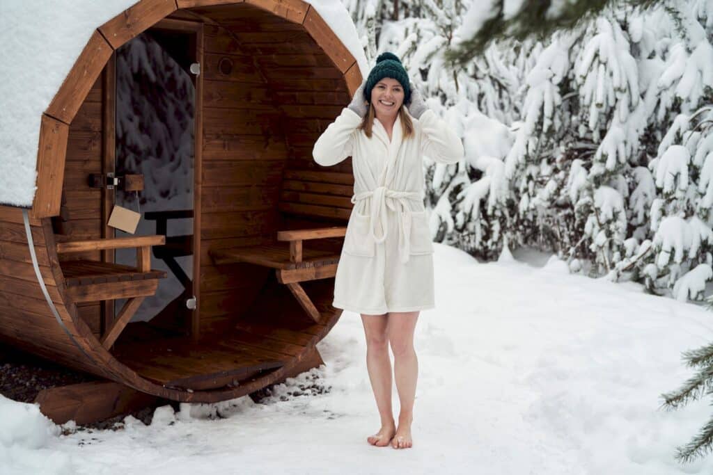 Une femme en peignoir blanc et bonnet vert se tient pieds nus dans la neige, souriante, devant un sauna en bois près d'un spa de nage en Suisse, entouré d'arbres enneigés.