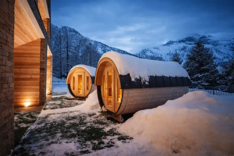Deux formes de sauna en bois avec des toits enneigés se trouvent à l'extérieur d'un bâtiment moderne au crépuscule, entourées de montagnes enneigées, leurs lumières chaudes s'allumant de manière invitante.