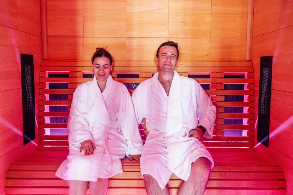 Deux personnes vêtues de peignoirs blancs sont assises et se détendent sur un banc en bois à l'intérieur d'un sauna très éclairé, baigné d'une lumière d'ambiance rose.