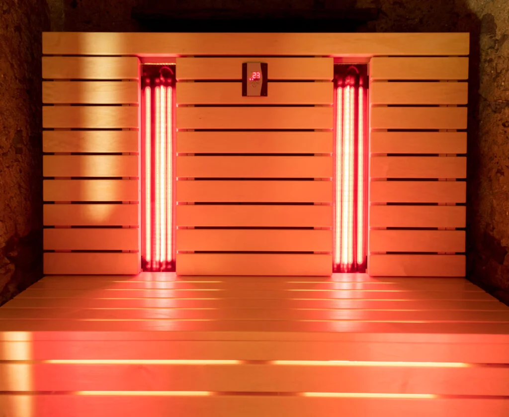 L'intérieur d'un sauna infrarouge en bois est baigné d'une lumière rouge provenant de deux radiateurs verticaux, et l'écran numérique affiche une température de 29 °C, créant ainsi une atmosphère chaleureuse et accueillante.