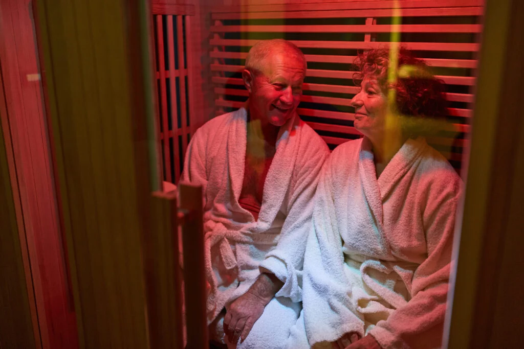 Un homme et une femme d'un certain âge, vêtus de peignoirs blancs, sont assis côte à côte et sourient dans un sauna baigné d'une lumière chaleureuse, profitant d'un moment de détente.