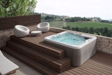 Une terrasse extérieure moderne dotée d'un jacuzzi encastré avec de l'eau claire et bouillonnante. Autour du bain à remous se trouvent d'élégantes chaises longues en osier blanc et une petite table. La terrasse donne sur une vue panoramique sur des vignobles verdoyants et des collines lointaines sous un ciel bleu clair.