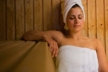 Une personne se détend dans un sauna, enveloppée dans une serviette blanche, les yeux fermés. Les murs et les bancs en bois créent une atmosphère chaleureuse. Une serviette enveloppe ses cheveux.