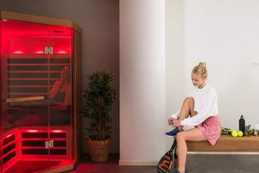 Une femme est assise sur un banc en bois dans un espace bien-être et attache ses chaussures. Derrière elle, une personne se détend dans un sauna infrarouge lambrissé de bois et éclairé en rouge. La pièce est décorée de plantes, de tableaux encadrés et d'une étagère contenant de petits objets.