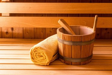 Un seau de sauna en bois avec une louche repose sur un banc en bois clair à côté d'une serviette jaune soigneusement roulée. L’arrière-plan présente des boiseries évoquant l’atmosphère chaleureuse et accueillante d’un sauna traditionnel.
