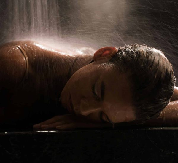 Une personne allongée sous un léger jet d’eau, comme dans un sauna tranquille. Le fond sombre accentue les gouttelettes d’eau tombant en cascade sur leur dos et leur tête, créant une atmosphère sereine et apaisante. Leurs yeux sont fermés et ils semblent détendus. sauna