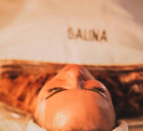 Une personne est allongée sur le dos, la tête enveloppée dans une serviette, les yeux fermés, à l’intérieur d’un espace étiqueté « Sauna ». L’éclairage est chaleureux, créant une atmosphère relaxante et sereine, illustrant parfaitement les bienfaits du sauna pour l’esprit et le corps. sauna