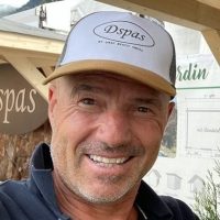 Un homme souriant portant une casquette de baseball blanche et beige avec le mot « Dspas » se tient à l’extérieur, peut-être sur un marché ou une foire, avec des structures en bois et une bannière en arrière-plan. Didier Deseneux