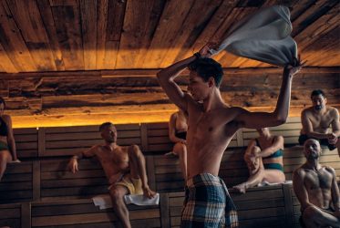 Une personne vêtue d'une serviette à carreaux agite un torchon dans un sauna en bois confortable, rappelant un bain nordique en Suisse. Plusieurs personnes sont assises sur des bancs en bois, certaines avec des serviettes. L'espace est doté d'un éclairage chaleureux et d'une atmosphère rustique.