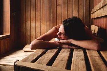 Une personne est allongée sur un banc en bois dans le sauna, la tête appuyée sur ses bras. La pièce est faite de boiseries et la lumière filtre par une fenêtre sur le côté gauche.