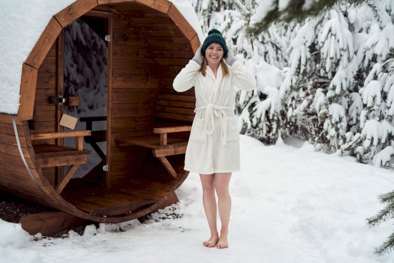 Une femme en peignoir blanc et bonnet vert se tient pieds nus dans la neige, souriante, devant un sauna en bois près d'un spa de nage en Suisse, entouré d'arbres enneigés.