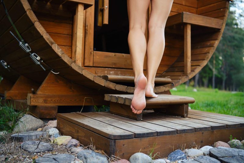 Une personne pieds nus monte des marches en bois et pénètre dans un sauna rond en bois en forme de tonneau, entouré de rochers et de verdure à l'extérieur - un moment de sérénité qui rappelle un spa de nage en Suisse.