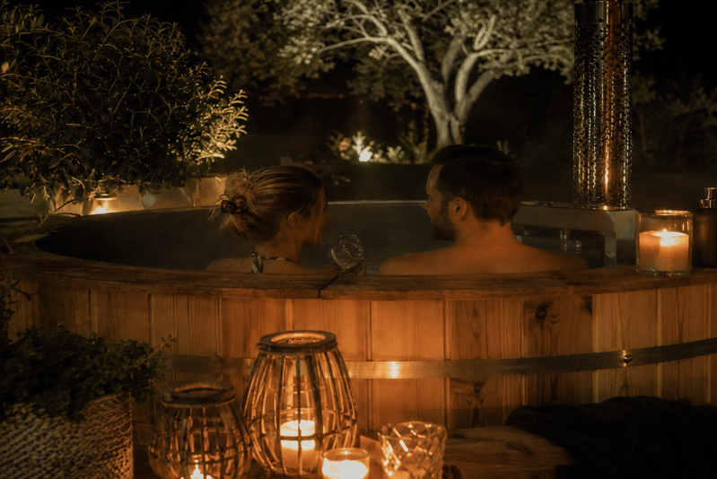 Un couple se détend dans un jacuzzi extérieur en bois, la nuit, entouré de bougies et de lanternes lumineuses, avec des arbres et un éclairage de jardin doux en arrière-plan.