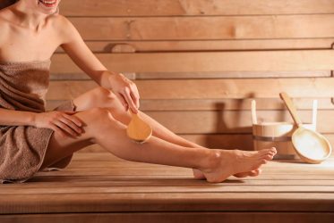 Une femme, vêtue d'une serviette, est assise sur un banc en bois de sauna ; elle sourit et se frotte la jambe avec une brosse en bois. Un seau et une louche de sauna se trouvent sur le banc à côté d'elle.