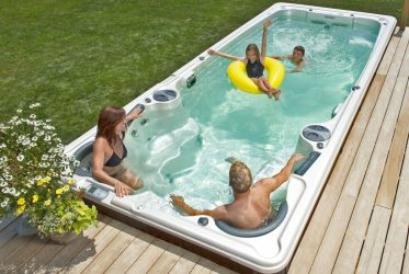 Une famille profite d’une journée ensoleillée dans un spa de nage dans la cour. Une femme en bikini est assise au bord, un homme se détend dans l’eau, un enfant flotte sur une chambre à air jaune avec les bras levés et un autre enfant nage à proximité. Les environs disposent d'une terrasse en bois et d'une pelouse verte.