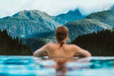 Une personne se détend dans une piscine, adossée au bord et contemplant une vue pittoresque sur les montagnes brumeuses et les forêts luxuriantes, rappelant l'ambiance tranquille que l'on trouve en Suisse. Le contraste serein entre l'eau claire et le paysage verdoyant offre une paix proche des expériences de sauna classiques.
