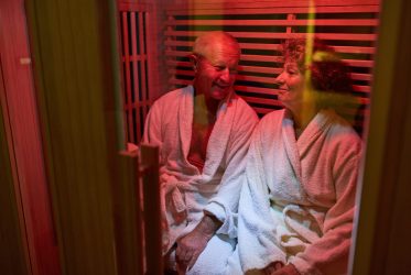Un homme et une femme d'un certain âge, vêtus de peignoirs blancs, sont assis côte à côte et sourient dans un sauna baigné d'une lumière chaleureuse, profitant d'un moment de détente.